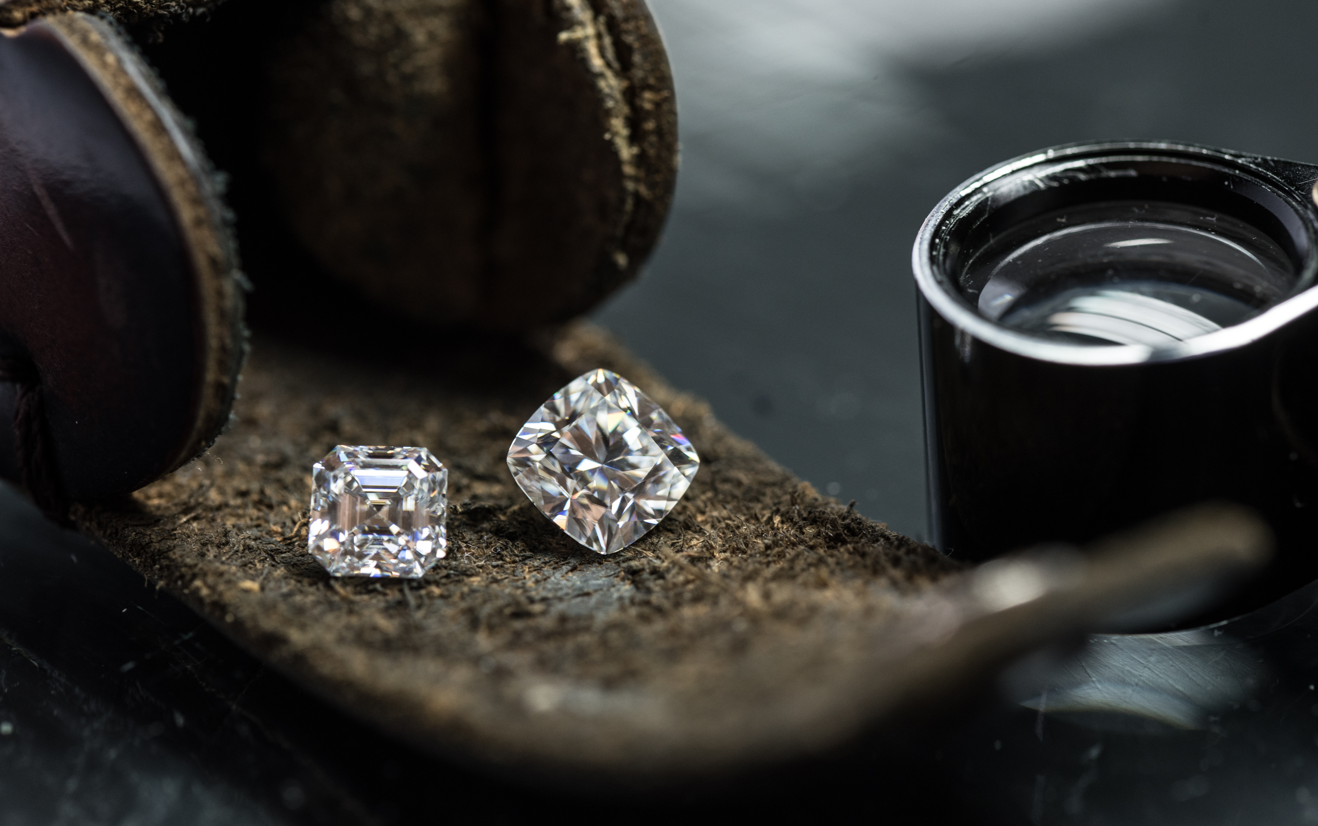 An asscher and a cushion-cut diamond on a leather parcel beside a jeweler's loupe